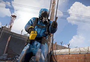 Een gewapende hazmat-medewerker in beschermende kleding, staande in een industriegebied met leidingen en constructies op de achtergrond.